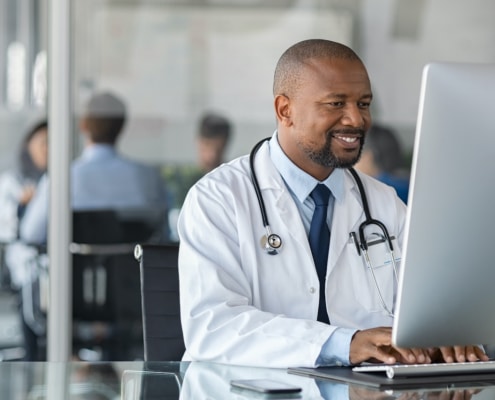 male doctor with a stethoscope around his neck smiles as he sits at a computer