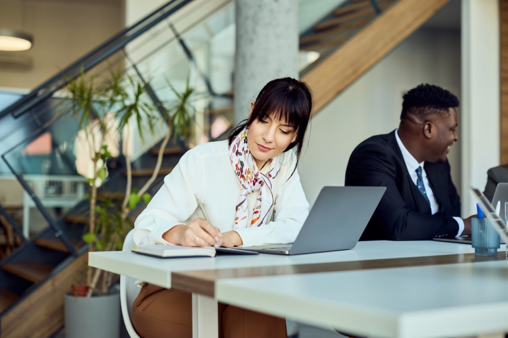 woman working in office writing something in notebook with her laptop on desk