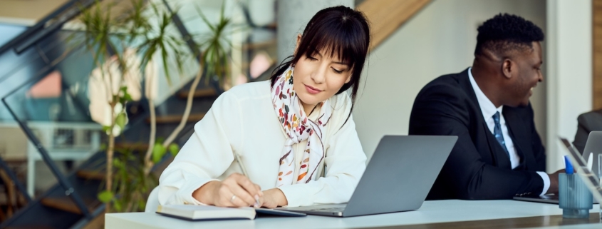 woman working in office writing something in notebook with her laptop on desk